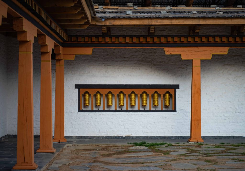 White wall with orange pillars and roof and several traditional golden Bhutanese prayer wheels in the wall